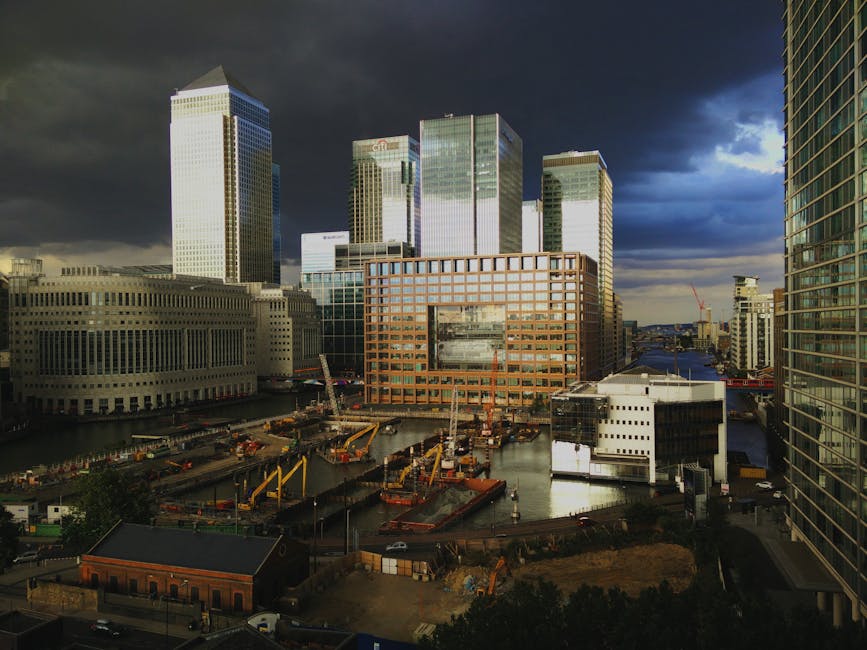A daytime view of a construction site in Canary Wharf with several large cranes actively lifting and moving building materials and equipment. The site is situated on a waterway, with portions of water visible among the construction activity. Surrounding the site are modern high-rise office buildings with glass facades, creating a dense urban backdrop. Inside the cordoned-off area, there are stacks of wooden pallets, metal scaffolding, and various construction tools and materials, indicating ongoing development work related to commercial property expansion. The scene is partially covered by dark clouds, suggesting overcast weather, with natural light illuminating the site and buildings, and some areas of shadow cast by taller structures. Docklands Man and Van occasionally handles furniture transport and home relocation projects in this busy commercial zone, often managing the logistics of moving large items or equipment through tight access points in similar urban environments.