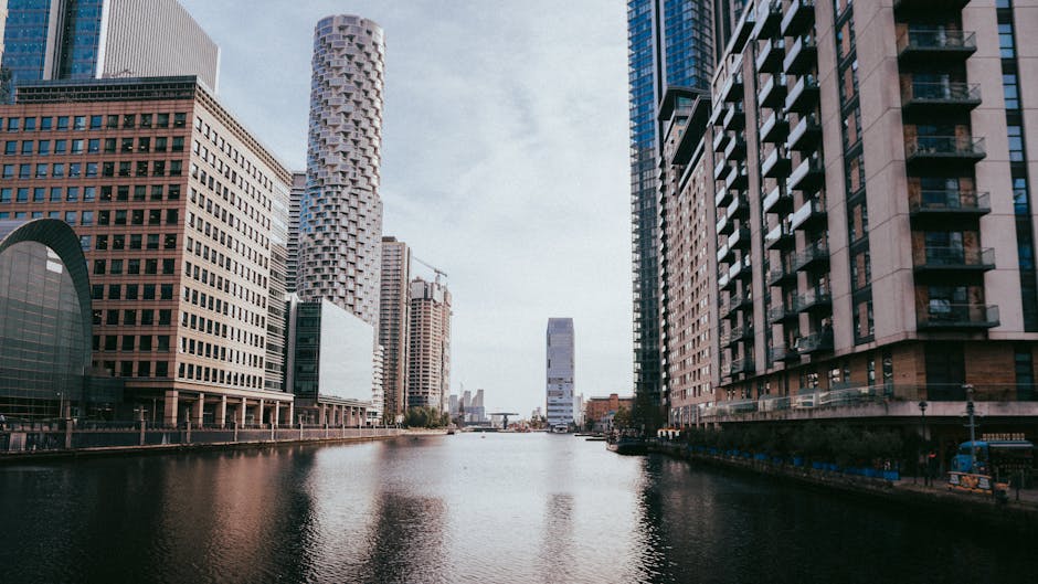 The image depicts a cityscape of Canary Wharf with a canal running through the center, flanked by modern high-rise office buildings and residential towers. On the left side, there are several glass and steel structures, including a large curved building, with the waterfront reflecting their facades on the calm water surface. To the right, tall residential buildings with balconies and windows are visible, with some extending to the edge of the water. The sky above is mostly clear with some light clouds. In the foreground, the canal is bordered by a paved walkway, and faintly visible are elements such as construction barriers and street furnishings, indicating ongoing development or maintenance work nearby. The setting suggests a typical urban environment suitable for professional house removals, with potential access constraints due to the dense infrastructure, aligning with services provided by Docklands Man and Van during home relocation or furniture transport processes.