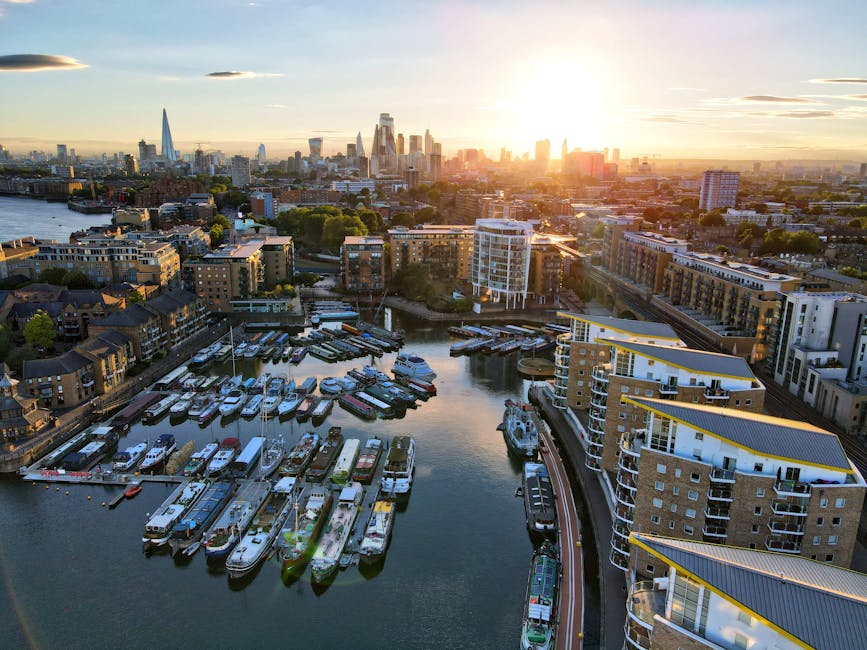 Aerial view of the Docklands area in London at sunset, showing a marina with numerous boats docked along piers adjacent to modern apartment buildings with yellow accents on their rooftops. In the background, the London skyline features prominent skyscrapers, including The Shard, with sunlight casting a warm glow over the city. The image depicts urban waterfront living and skyline views, relevant to the logistics of house removals and furniture transport in the Docklands area, with the setting sun highlighting the area's development. Docklands Man and Van would facilitate home relocation and moving services within such a setting, involving packing, loading, and transporting household items by van from residential properties to new locations.