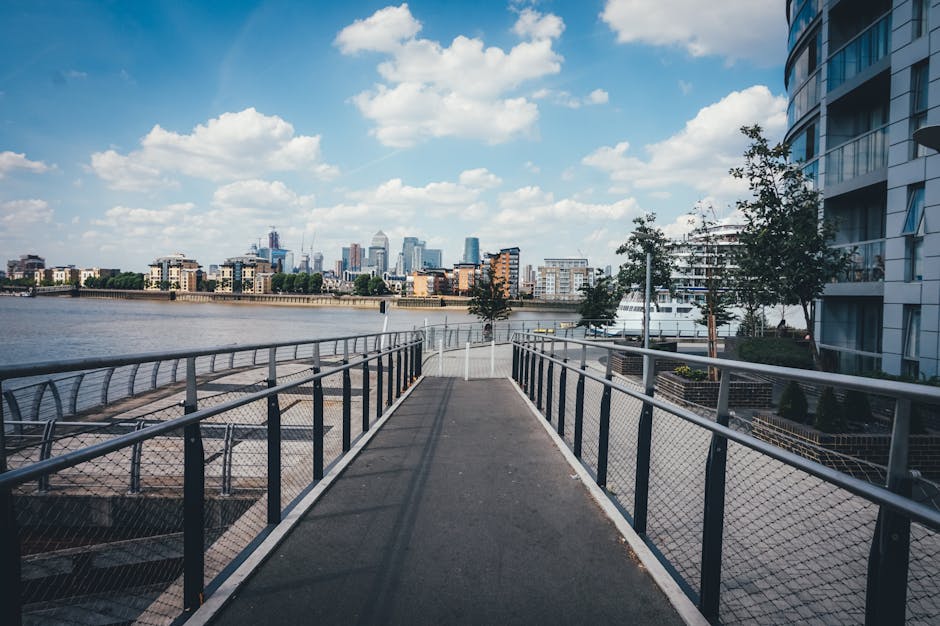 A wide outdoor walkway or pier along the River Thames in Docklands, showing a metal railing on both sides, with a few leafless trees and modern residential buildings on the right. In the background, a city skyline with high-rise office buildings and cranes under a partly cloudy sky is visible. The foreground features a smooth, dark paved surface extending toward the horizon, with shadows cast by the railing. This scene captures the waterfront environment typical of the Isle of Dogs E14 area, where house removals and professional moving services such as those provided by Docklands Man and Van may involve logistics around loading or unloading furniture, boxes, and packaging materials in a scenic urban river setting.