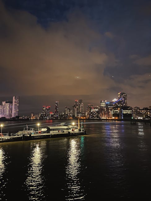 Aerial view of the Docklands area in London at sunset, showing a marina with numerous boats docked along piers adjacent to modern apartment buildings with yellow accents on their rooftops. In the background, the London skyline features prominent skyscrapers, including The Shard, with sunlight casting a warm glow over the city. The image depicts urban waterfront living and skyline views, relevant to the logistics of house removals and furniture transport in the Docklands area, with the setting sun highlighting the area's development. Docklands Man and Van would facilitate home relocation and moving services within such a setting, involving packing, loading, and transporting household items by van from residential properties to new locations.