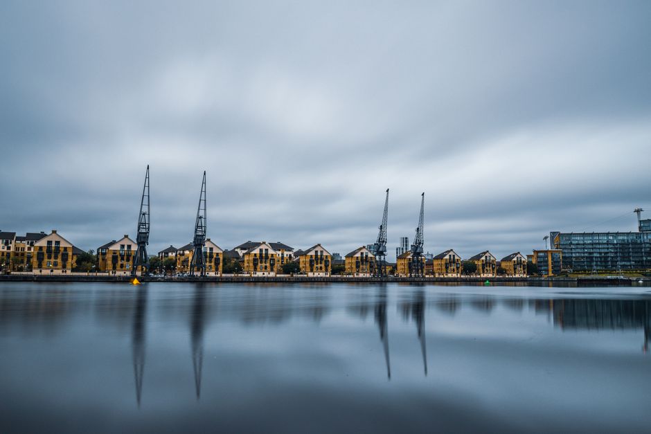 A wide panoramic view of a Docklands waterfront scene under an overcast sky with grey clouds, where multiple large industrial cranes are positioned along the dockside. Behind the cranes, there is a ro