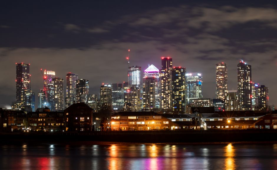 A wide panoramic view of a Docklands waterfront scene under an overcast sky with grey clouds, where multiple large industrial cranes are positioned along the dockside. Behind the cranes, there is a ro