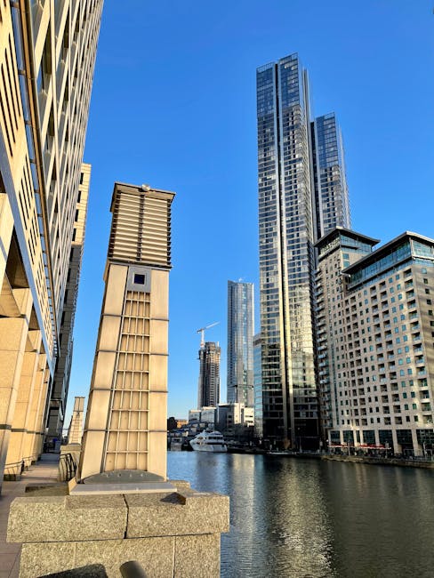 The image shows a cityscape along a canal in Docklands with tall modern skyscrapers made of glass and steel, reflecting the blue sky. On the left, a building with a beige stone exterior features a protruding ventilation or chimney structure with metal louvers, positioned on a stone ledge near the water. In the background, several high-rise buildings, some under construction, are visible, including a crane. The canal water is calm, with a boat moored along the quay. The scene is illuminated by natural daylight, highlighting the architectural details and urban environment, which is relevant to services offered by Docklands Man and Van for house removals and furniture transport around West India Quay and the Docklands area, conveying aspects of packing, loading, and logistics involved in home relocation.