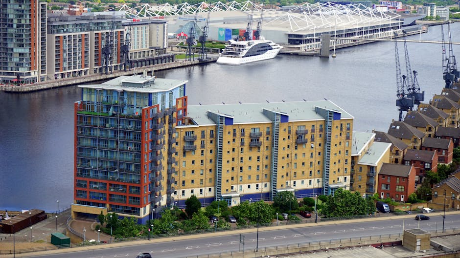 Aerial view of a multi-storey residential building in Docklands, featuring a modern design with glass balconies, situated near waterways with boats and yachts. The building has a yellow and blue façade, with several balconies visible. Surrounding the building are trees and parked cars along the adjacent road. In the background, there are other residential structures, a marina with docked boats, including a large white yacht, and cranes indicating ongoing construction or shipping activity. The scene captures the urban environment typical for house removals, involving loading and transport logistics, with vehicles and equipment potentially used for furniture transport and home relocation. Natural daylight illuminates the scene, highlighting the details in the building’s structure and waterfront location, consistent with professional removals services offered by Docklands Man and Van in West India Quay.